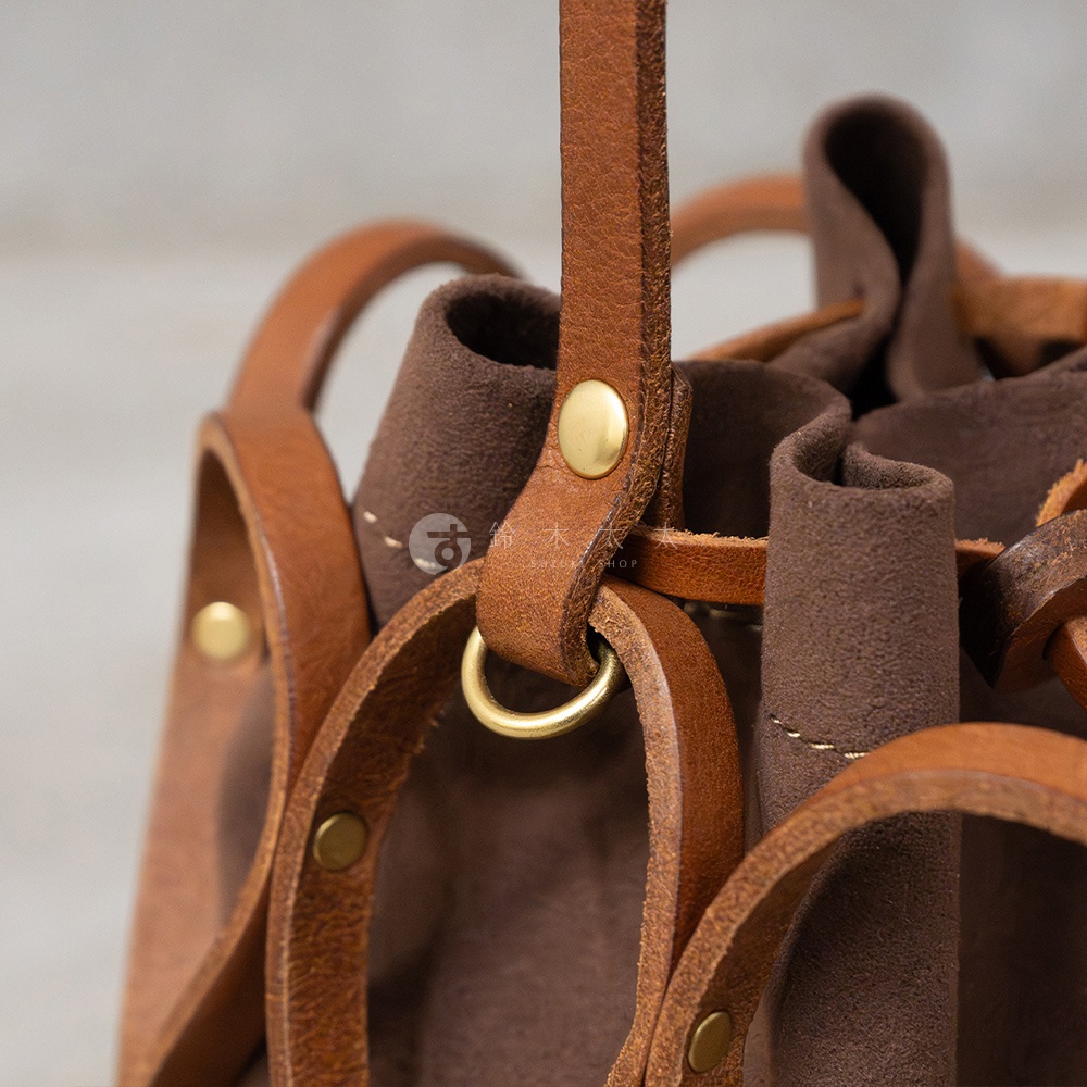 細膩的HERZ Organ編織水桶包，展現職人手作皮革工藝，駝色和棕色皮革的溫潤質感，搭配金色五金細節，透過局部特寫呈現精緻的縫線與織帶細節。