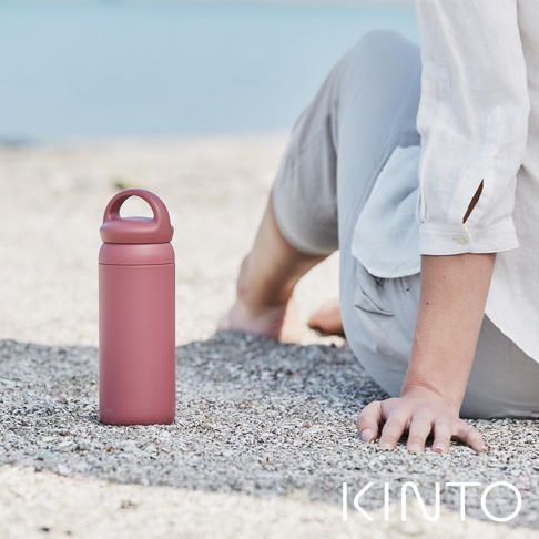 一個玫瑰色、帶提把的保溫瓶，瓶身呈現霧面質感，瓶蓋與瓶身同色，放置於沙灘上。