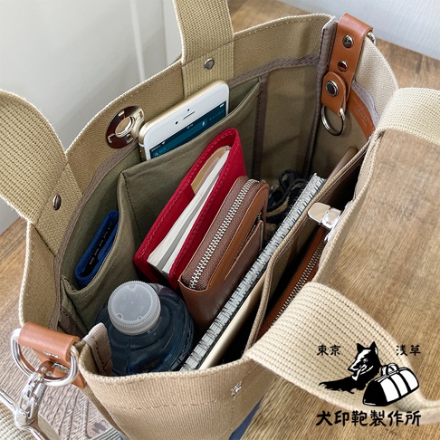開放式帆布托特包，內有手機、紅色筆記本、咖啡色皮夾、筆記本和水瓶，包包外部有品牌標誌，材質為純棉帆布。