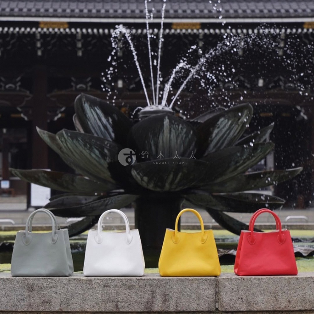 四款手提托特包，分別為灰色、白色、黃色與紅色，皆為皮革材質，造型簡約，在日式建築與蓮花噴泉前展示。