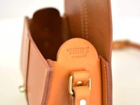 職人手作皮革皮扣單肩包，採用天然皮革材質，呈現溫潤的棕褐色調，帶有精緻的縫線與金屬皮扣細節，中央壓印有「HERZ」品牌標誌與「MADE IN JAPAN」字樣。