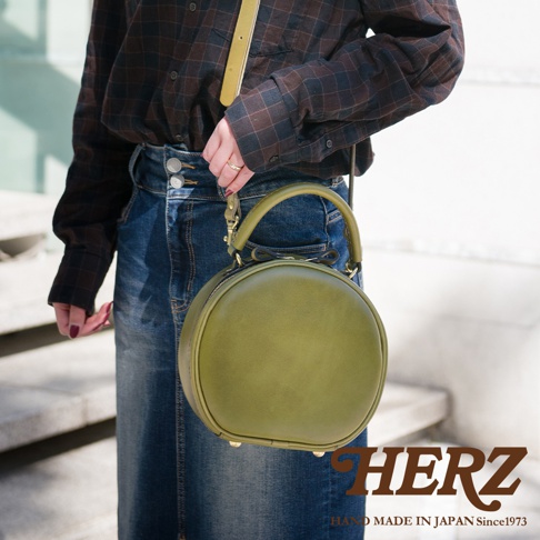 日本HERZ品牌，職人手作皮革兩用圓形包，綠色皮革搭配牛仔裙，包上印有HERZ品牌字樣。