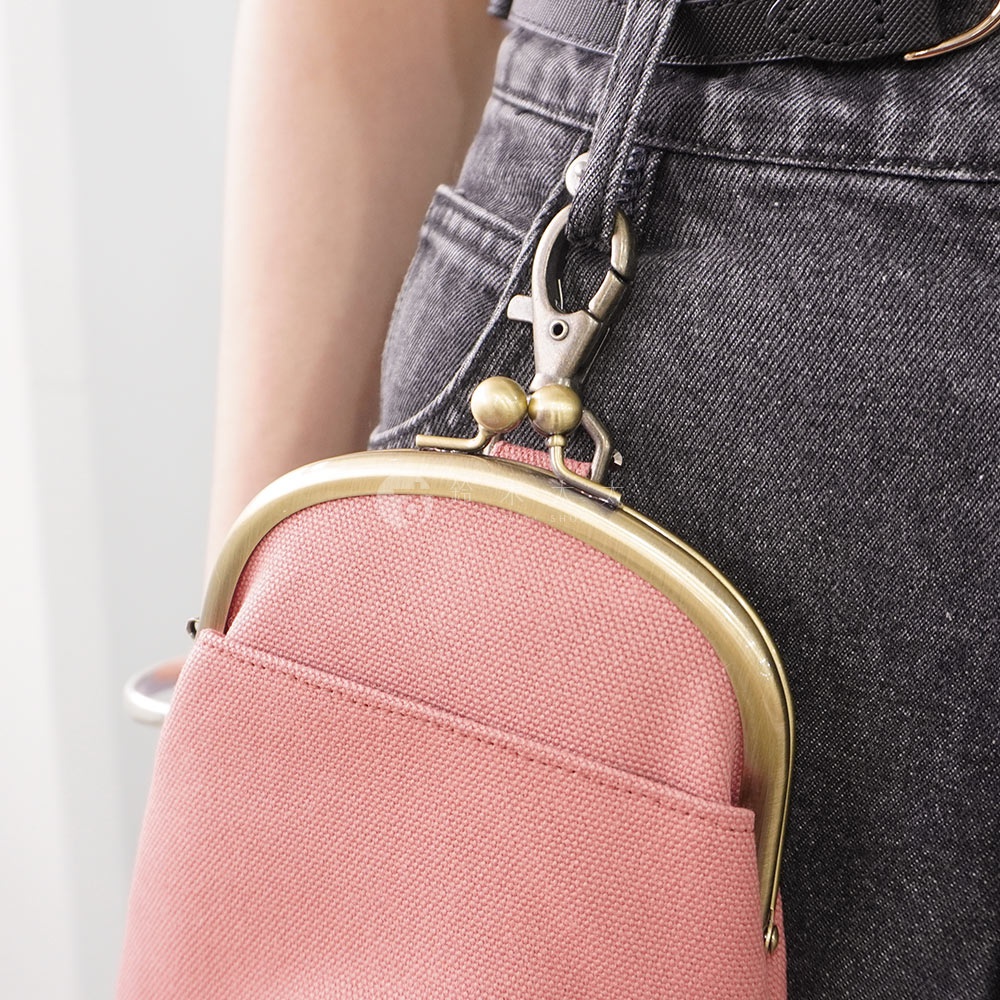 側腰掛著一個粉紅色帆布質地的口金包，包包的開合處是金色的金屬框架，上面有一個金屬掛鉤，掛鉤連接到褲子上的鈕扣。