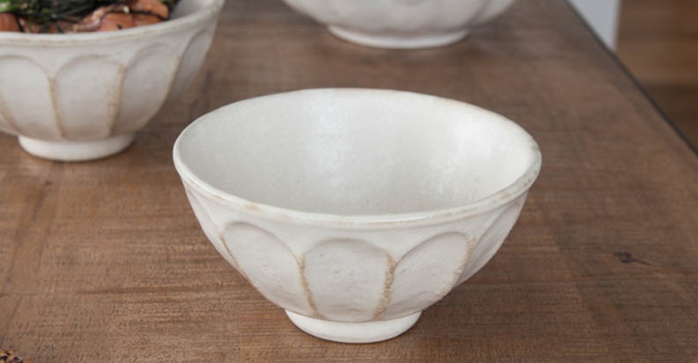一個白色陶瓷丼碗，碗身帶有粉引紋理及六瓣花紋，底部有圓形圈足，置於木質桌面。