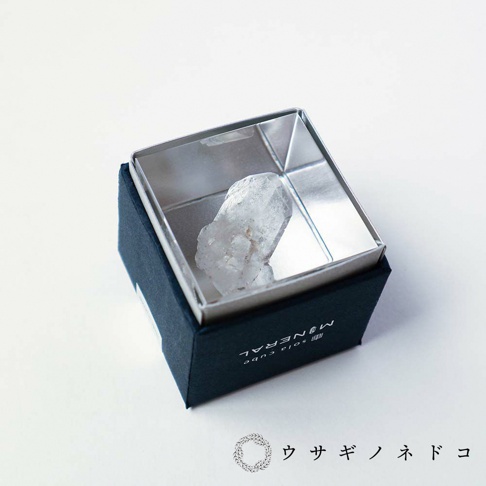 深藍色盒子內，一個銀色金屬框中，陳列著一塊半透明的白色水晶礦石，其表面呈現不規則的晶體紋理。