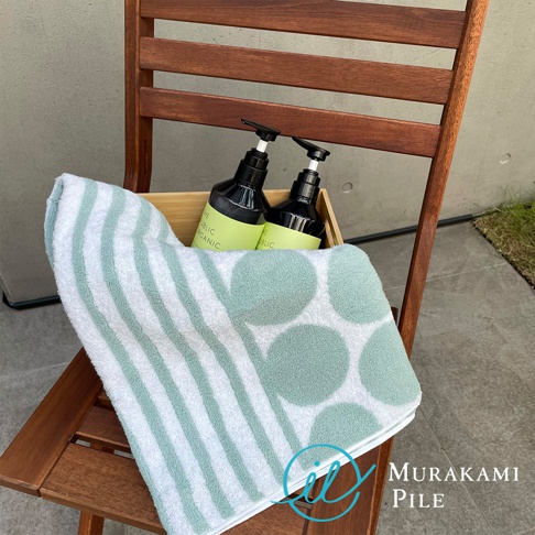 一座木質椅子上擺放著一條印有綠色圓點和條紋的毛巾，毛巾旁邊的木盒裡有兩瓶綠色標籤的洗浴用品，右下角有 Murakami Pile 的文字。