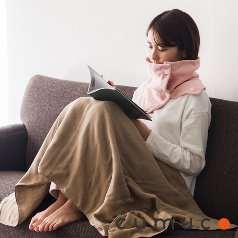 一名女子坐在沙發上，膝蓋上蓋著一條棕色毯子，膝蓋上放著一本翻開的書，她脖子上圍著一條粉紅色保暖頸套，脖套上有兩顆白色鈕扣，女子左側的角落印有「cumoco」的黑色字樣，右側角落有一個橙色圓形標誌。