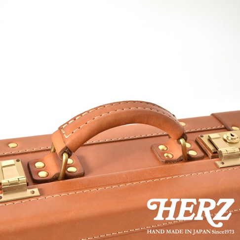 職人手作的淺棕色皮革手提行李箱，帶有黃銅色五金配件，包含提把、鎖扣和鉚釘，縫線清晰，展現英倫風格。