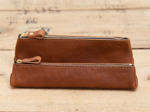 兩個棕色皮革製成的三角收納筆袋，帶有金色拉鍊和皮革拉環，其中一個較大，另一個較小，皆放置於木紋背景上。