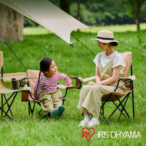 兩張卡其色露營摺疊野餐椅，配備置物網袋和杯架，椅子框架為黑色金屬。一位母親和孩子坐在椅子上，背景為綠草地和遮陽布。