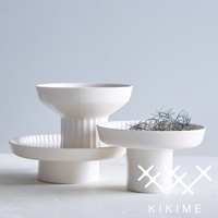 三款白色陶瓷高腳平盤，其中左側為較高、帶有垂直溝槽的盤子，中間為較扁平、帶有放射狀紋路的盤子，右側盤中飾有綠色細長植物，盤緣帶有波浪狀紋路。