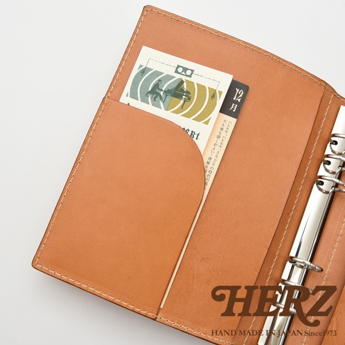 職人手作皮革A5手帳/記事本書套，淺棕色皮革，內有卡片夾層，置入一張印有鋼琴圖案的卡片，以及寫有日文的紙張。書套內側有銀色六環活頁夾。