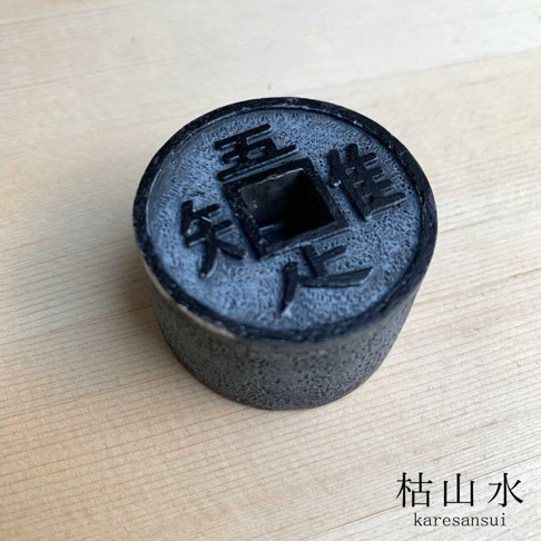 這是一個呈現出仿古銅錢外觀的黑色圓柱體擺飾，頂部刻有五、唯、知、足四個漢字，中間為正方形孔洞，整體具有岩石般的顆粒質感，放置在淺木色的桌面上，右下角標註有枯山水 karesansui 字樣。
