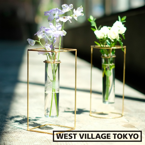 兩個黃銅圓柱玻璃花器，前方花器內插有紫白色小碎花，後方花器內插有白色玫瑰，皆置於簡約的黃銅方形支架中，呈現淡雅的居家佈置風格。