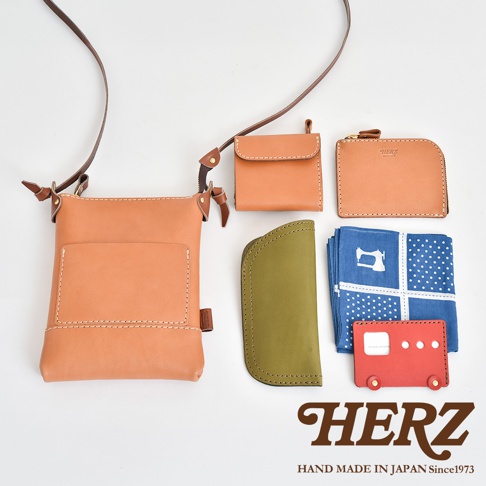 職人手作皮革直式隨身單肩包，搭配同色系皮革零錢包、長夾，以及橄欖綠皮革收納袋、藍色印花手帕與紅色造型卡夾，展現精緻工藝。