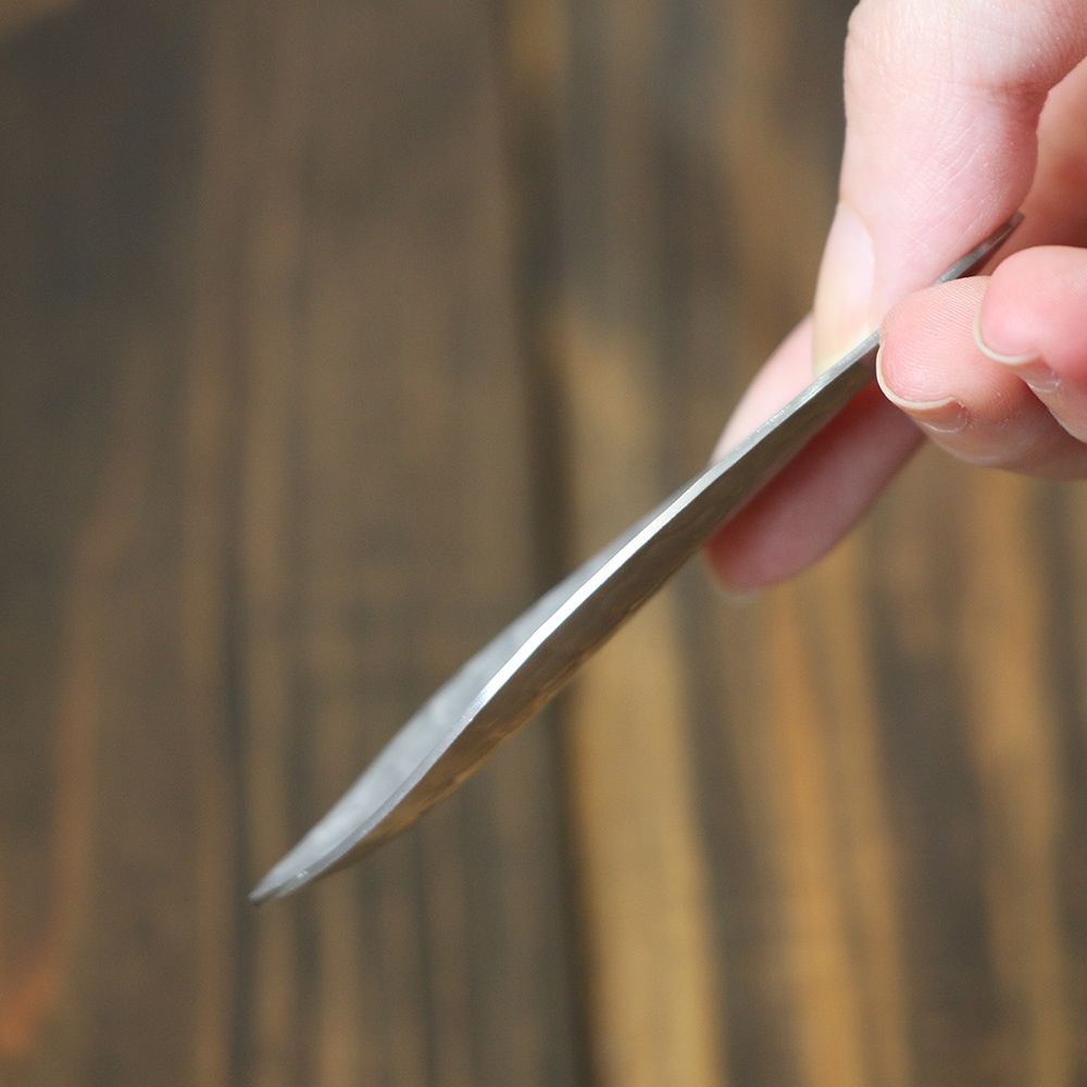 一只手拿著一把銀色不鏽鋼茶勺，茶勺呈現細長的彎曲形狀，勺頭部分較為扁平，背景為深棕色木紋。