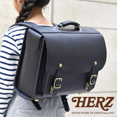 黑色皮革橫式書包，配有金屬扣環和提把，由一位穿著條紋上衣的小女孩背著。