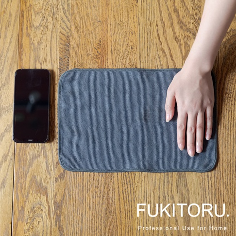 深灰色超細纖維擦拭布，帶有細緻的條紋紋理，放置於木質桌面，一旁為黑色手機，右手置於布上。