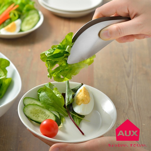 一只手使用不沾桌調理幫手夾夾取生菜，生菜葉片翠綠，夾子為金屬材質，帶有弧形設計，正將生菜放入裝有小番茄、小黃瓜片和水煮蛋的白色盤中。