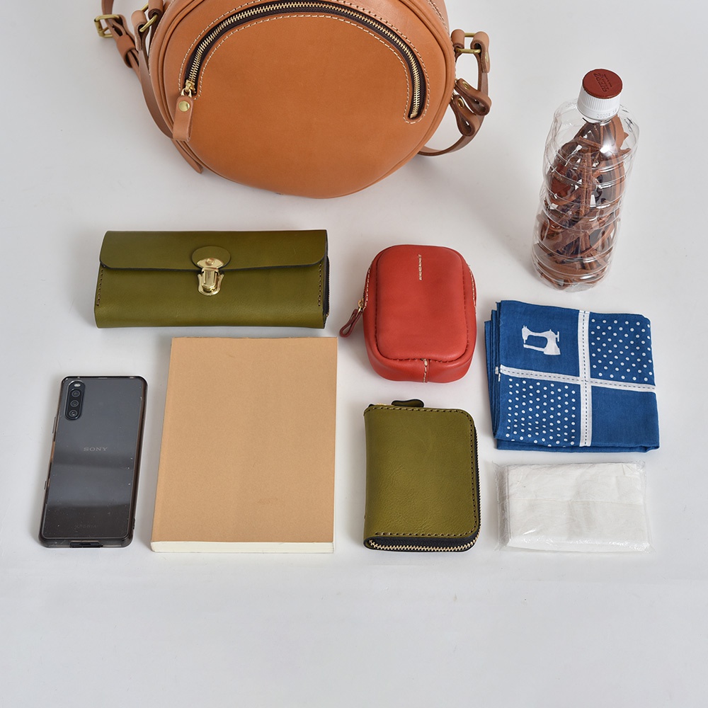 職人手作皮革小圓包 CK-19 M，展示了包含棕色皮革圓包、綠色長夾、紅色小包、綠色零錢包、筆記本、手機、藍色印花手帕、紙巾和一瓶水的擺設。