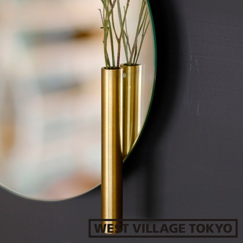 黃銅色金屬材質的壁掛式雙管花器，插入綠色植物的細長莖幹，花器固定於一面圓形鏡面後方，鏡面反射出模糊的背景。