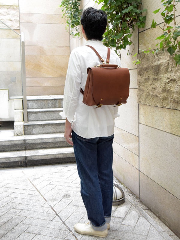一只棕色皮革雙扣兩用側背包，揹在一位穿著白色襯衫和牛仔褲的人身上。