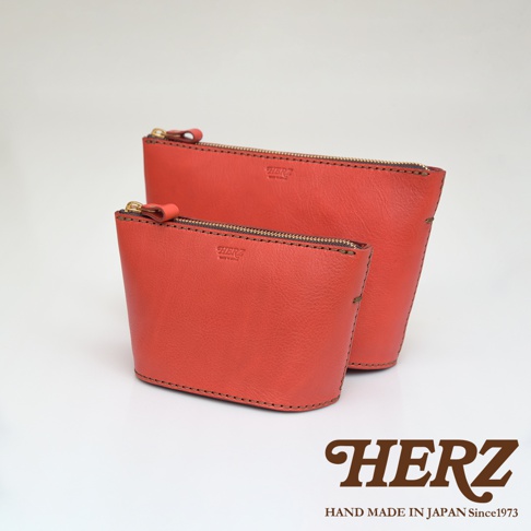 兩個橘紅色皮革製的職人手作三角收納包，一前一後擺放，材質呈現細緻皮革紋理，並配有金色拉鍊。