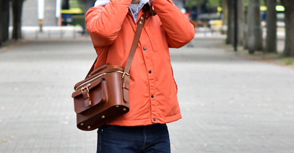 一名男子身穿橘色夾克，肩上背著一個棕色皮革相機包，站在一條鋪滿落葉的步道上。