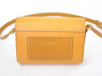 職人手作皮革寬扣帶馬鞍包，以天然皮革製成，呈現溫潤的焦糖色。包款設計簡約，正面有一個方形外袋，邊緣有精緻的縫線裝飾。寬版的皮革背帶透過金屬環連接包身。