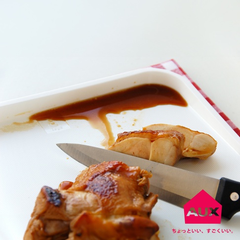 白色防溢大砧板上，擺放著切塊的烤雞肉，部分肉塊淋有醬汁。一支不鏽鋼刀正切著雞肉，刀柄為黑色。背景有一塊紅白色格紋布。