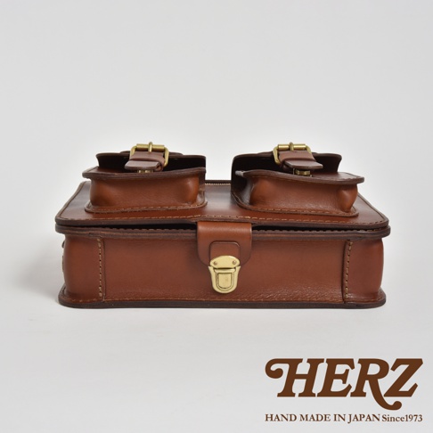 職人手作皮革雙口袋兩用單肩包 A-19，採用咖啡色皮革製作，包身有兩個上方帶有皮帶釦的翻蓋口袋，正面有一個黃銅色金屬鎖扣，整體呈現簡約復古風格。