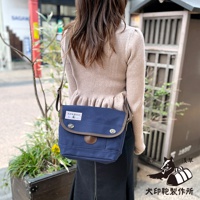 一名女性肩背著一個深藍色純棉帆布郵差包，包蓋上有棕色皮革飾邊和兩個銀色金屬扣。