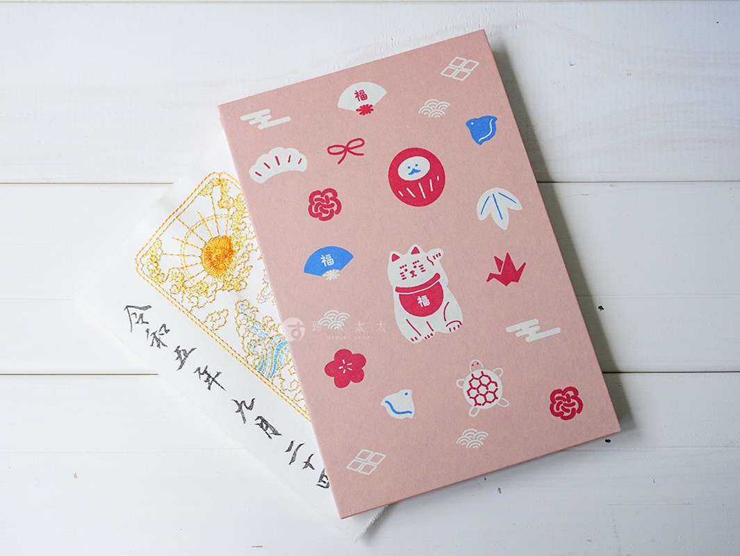 粉紅色和紙御朱印帳，印有招財貓、達摩、山茶花、扇子、千羽鶴、烏龜等可愛圖案，另有一張白色布質御朱印，繡有金色太陽與雲朵圖案，字跡為黑色。
