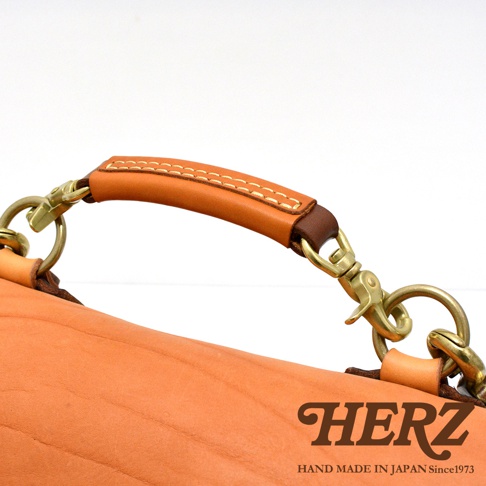 職人手作皮革鹿角扣繩郵差包，特寫皮革提把與黃銅五金扣環，包身為淺棕色皮革，提把處縫線明顯。