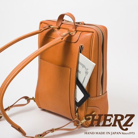 一個棕褐色的皮革後背包，有著精緻的縫線細節，背包右側有一個拉鍊口袋，內有筆記本和卡夾。