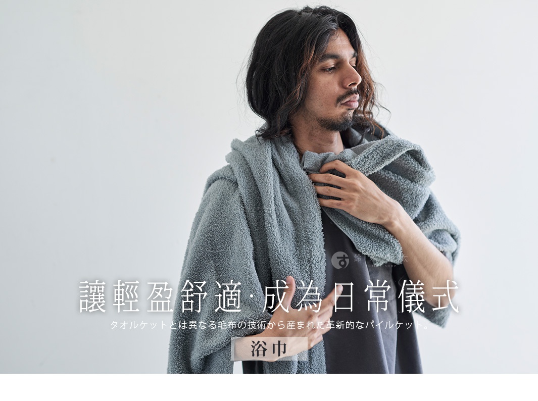 一位年輕男性穿著一件灰色的蓬鬆毛巾，他將毛巾披掛在肩膀上，左手輕撫胸前的毛巾。背景是純白色，營造出簡潔的氛圍。圖片下方有中文和日文的標語，標語內容大致為「輕盈舒適，成為日常儀式」以及「浴巾」。