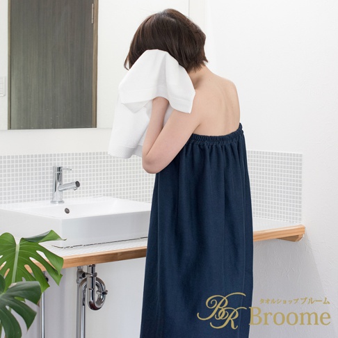 一位女性穿著深藍色、鬆緊帶平口浴裙，正在用白色毛巾擦臉。浴裙材質為泉州棉紗混紡，質感柔軟。