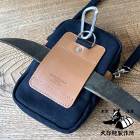 深藍色石洗帆布斜背手機包，附有棕色皮革吊牌，吊牌上印有品牌名稱與產地。包包配備銀色登山扣和可調節肩帶。