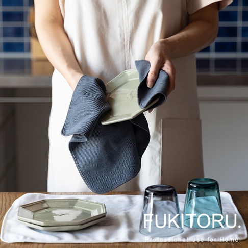 兩塊深灰色的超極細纖維擦拭布，其中一塊正在擦拭一個淺綠色的六角形盤子。桌面上另有兩個疊放的六角形盤子和兩個玻璃杯。