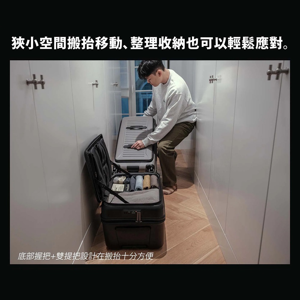 一名男子在狹窄的走廊空間中，正將一個黑色與灰色相間的 AZPAC 前開口胖胖行李箱打開進行收納，行李箱底部與側邊配有方便搬運的提把設計，即使在狹小環境中也能輕鬆整理物品。