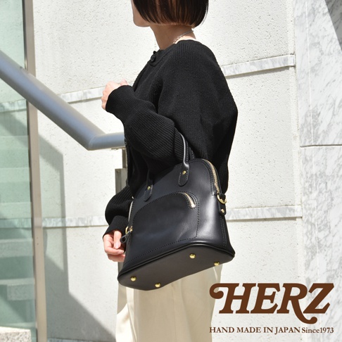 一位女性手持黑色經典貝殼造型皮革手提包，包身光滑，設有金色拉鍊和金屬腳釘。