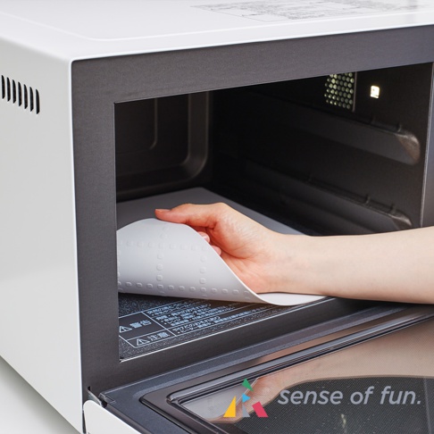 一位女士的手正在將一個圓形的、帶有圓點圖案的白色耐高溫微波爐墊放入一個白色微波爐的內部，墊子下方有日文警告標示。