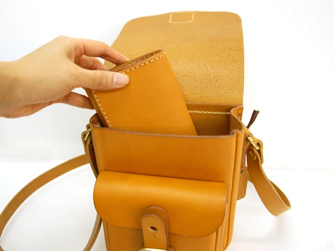 職人手作的直式單肩包，採用天然皮革材質，呈現溫潤的棕褐色。包身結構清晰，帶有皮扣翻蓋設計，並展示一個手正將皮夾放入包內。