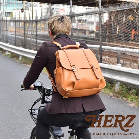 一位背著日本HERZ棕色皮革後背包的人正騎乘腳踏車，包款設計採用經典雙扣開合與屋瓦風格頂部結構，展現職人手作的質感，背景為戶外道路與鐵軌圍籬。
