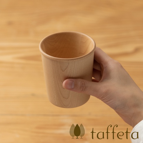 一隻手拿著一個淺木色楓木寬口杯，杯身呈現自然的木紋，杯緣圓潤。