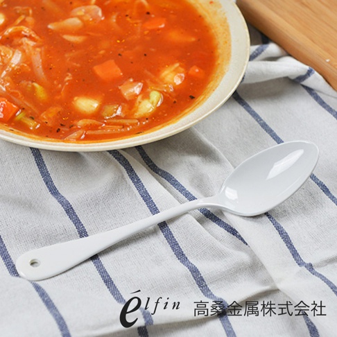 一隻日製白琺瑯餐匙，湯匙的柄上有個小圓孔，放置於藍白條紋的布上，旁邊有一碗番茄蔬菜湯。