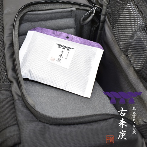 一個白色帶有紫色壓紋紙包裝的古來炭迷你調濕木炭，置於黑色背包的內袋中。包裝上印有日文文字及圖案。