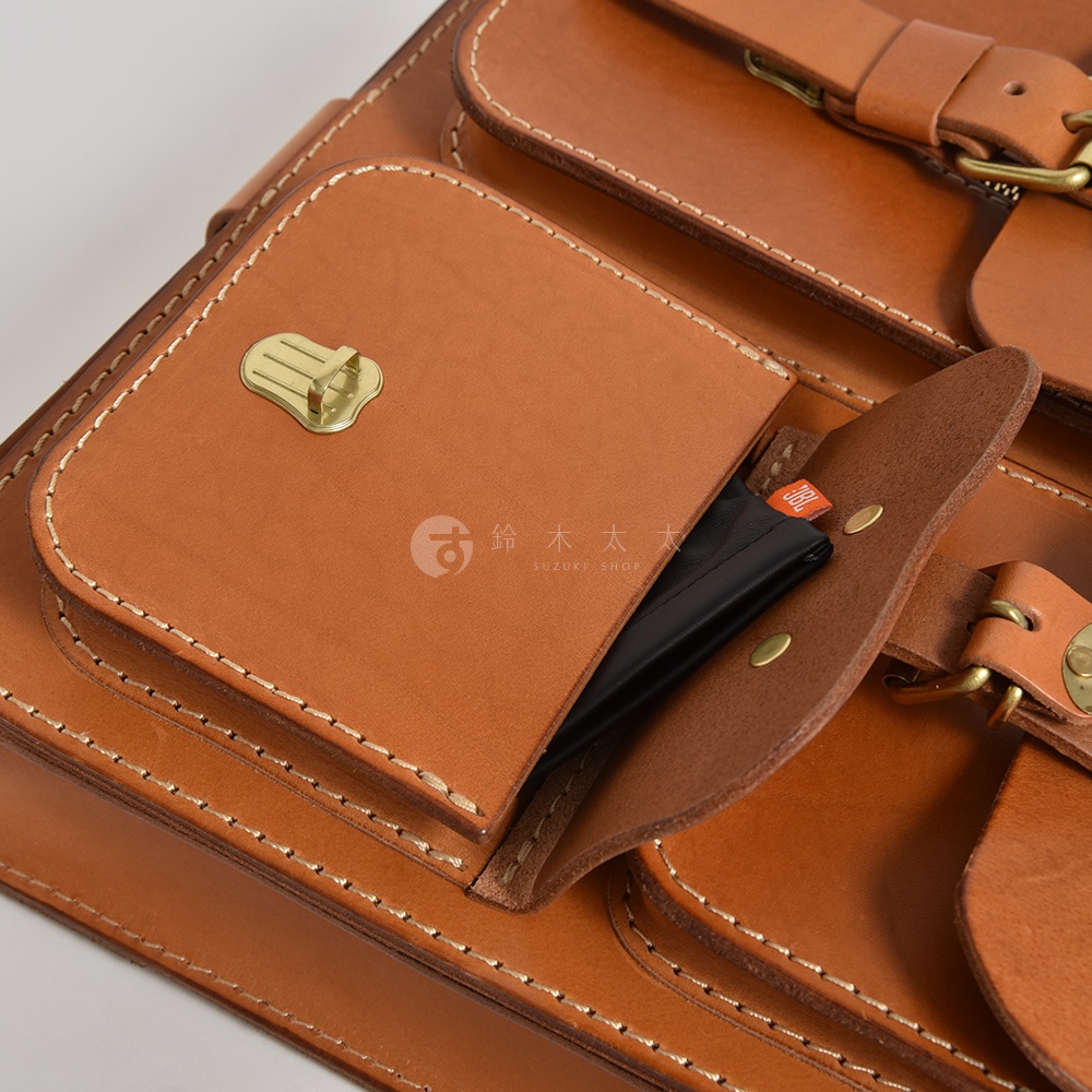 職人手作皮革三口袋三用後背包，採用淺棕色天然皮革，車線清晰。包款正面有兩個帶有黃銅扣具的口袋，其中一個口袋露出部分黑色物體。