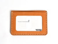 職人手作皮革橫式票券收納夾，採用天然鞣製皮革，呈現溫潤的橘棕色調，邊緣以米白色縫線工整車縫。正面中央設有透明PVC票券夾層，內嵌「The Heart of Craftsmanship」字樣與小人圖標，右下角壓印黑色「HERZ」品牌標誌。