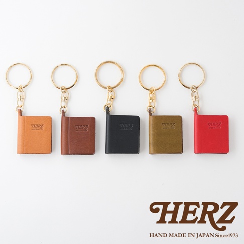 五款職人手作皮革書本造型鑰匙圈，包含橘棕色、深棕色、黑色、橄欖綠和紅色，皆有金色金屬環，皮革材質，印有HERZ品牌標誌。